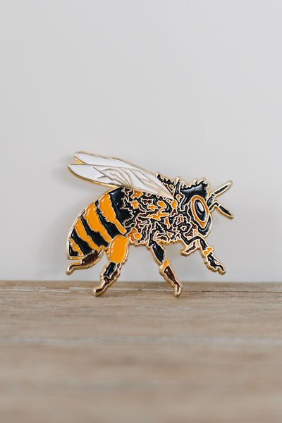 Custom hand-drawn honeybee enamel pin with gold detailing, black and orange stripes, and white wings, displayed on a neutral white background.