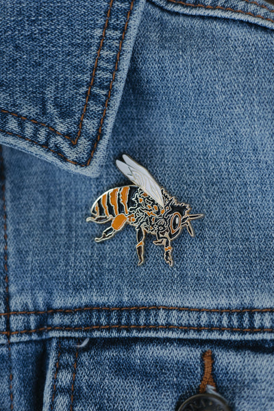 Custom hand-drawn honeybee enamel pin with gold detailing, black and orange stripes, and white wings, displayed on a denim surface against a neutral background.