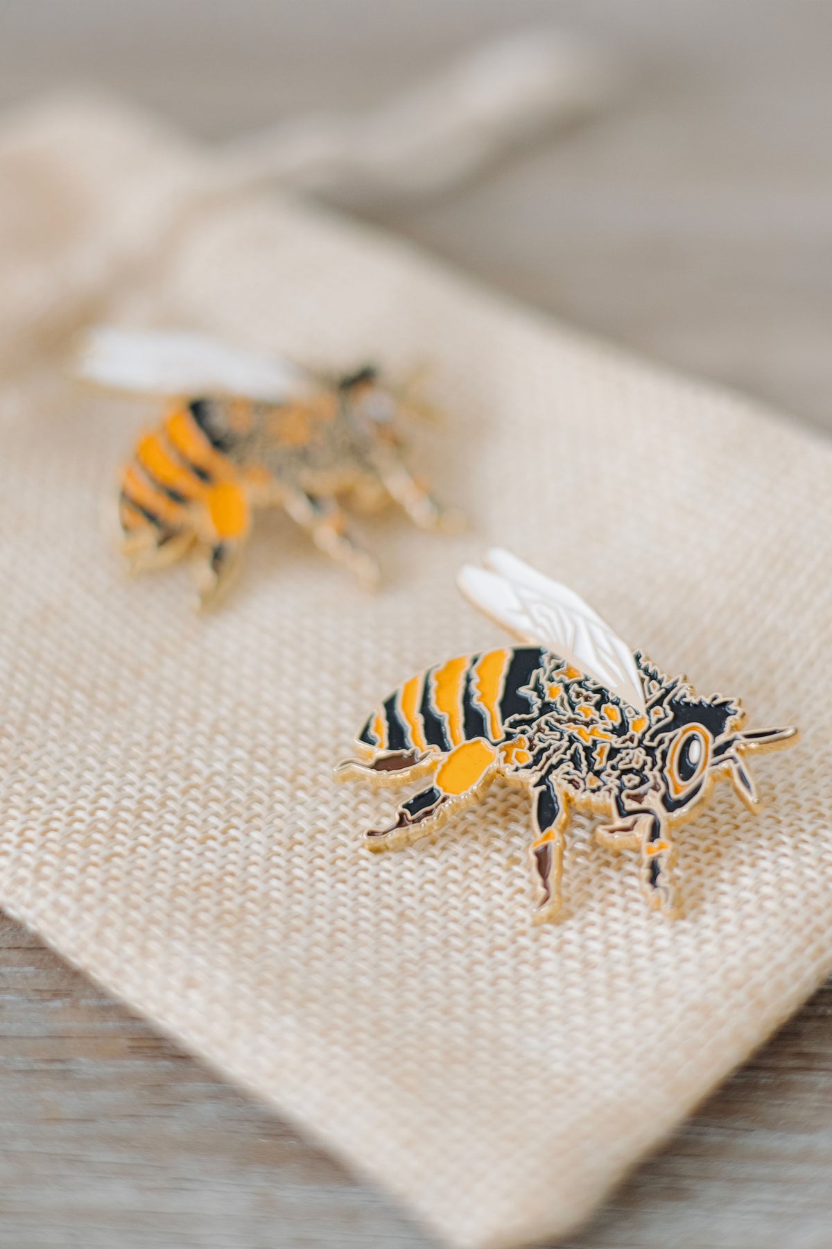 Custom hand-drawn honeybee enamel pin with gold detailing, black and orange stripes, and white wings, displayed on a linen towel surface against a neutral background.