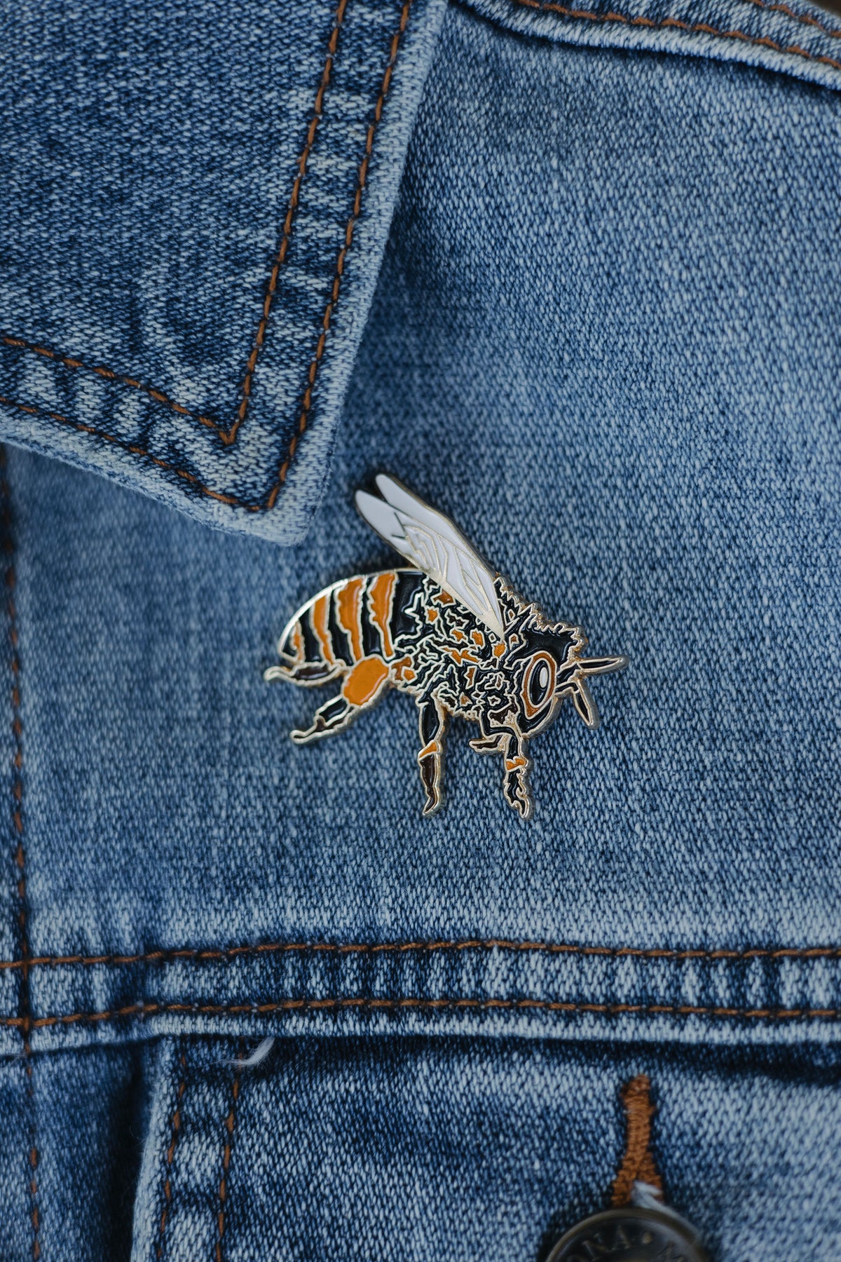 Custom hand-drawn honeybee enamel pin with gold detailing, black and orange stripes, and white wings, displayed on a denim surface against a neutral background.