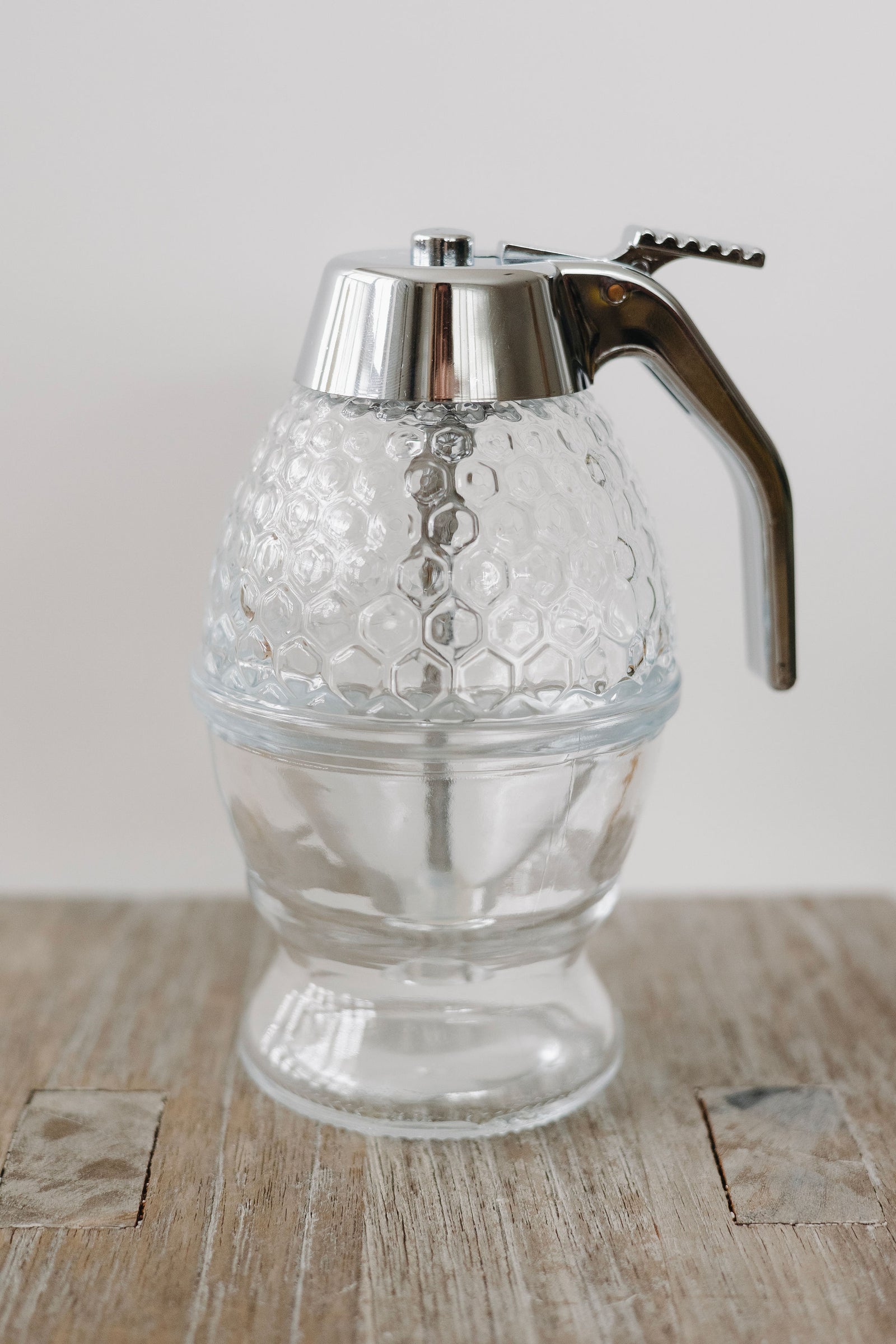 Glass honey dispenser with honeycomb texture filled with golden honey, placed on a wooden surface with a striped towel and small white flowers.