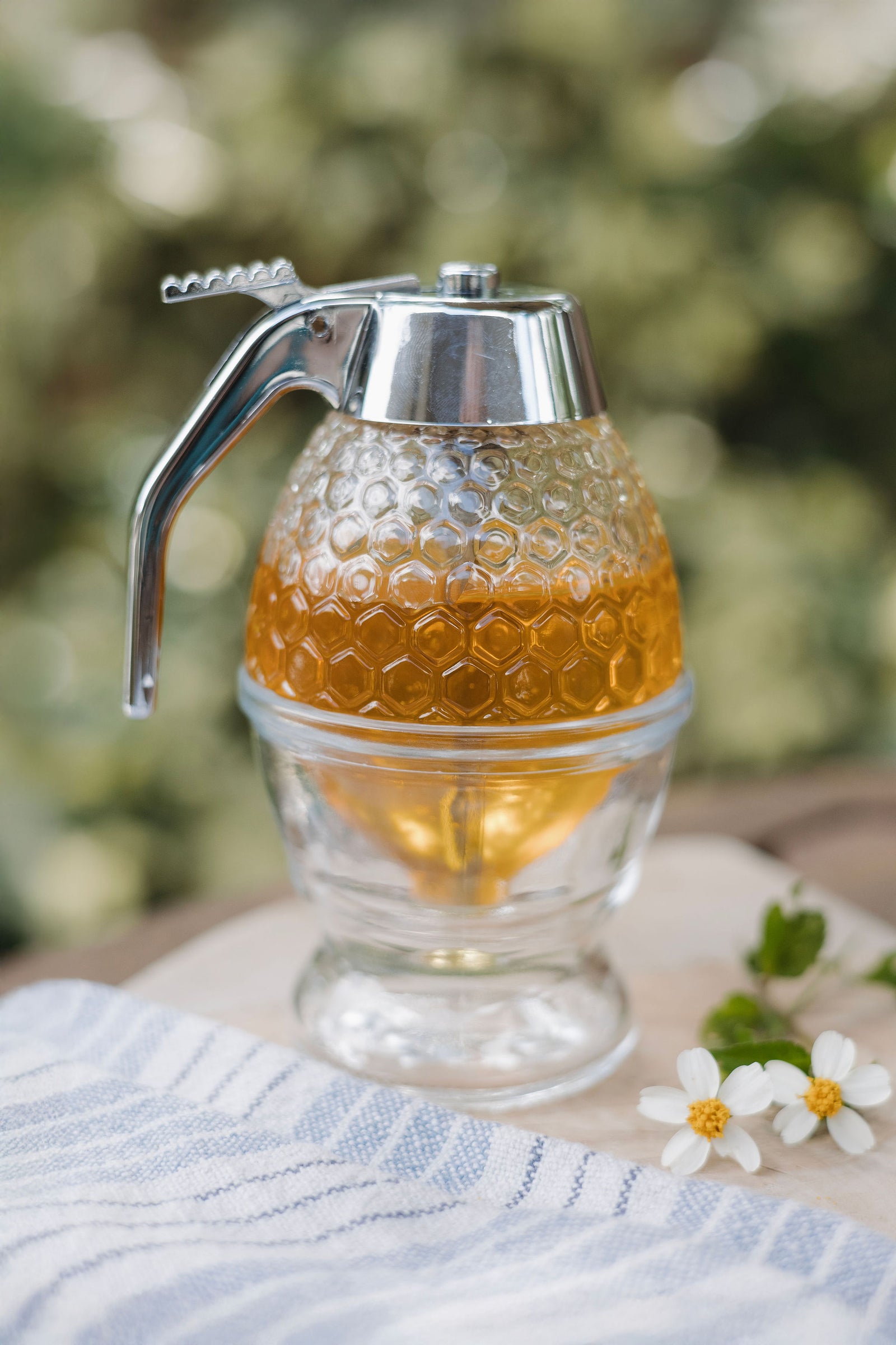 Glass honey dispenser with honeycomb texture filled with golden honey, placed on a wooden surface with a striped towel and small white flowers.