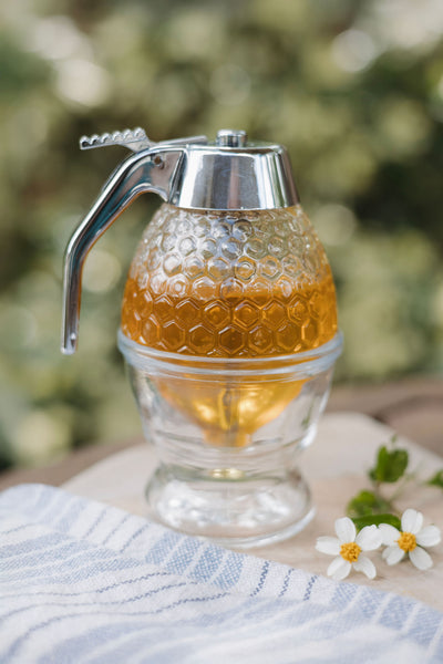 Glass honey dispenser with honeycomb texture filled with golden honey, placed on a wooden surface with a striped towel and small white flowers.